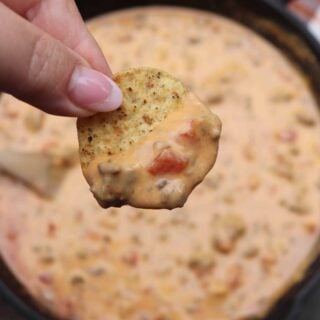 Chip with rotel dip being held over a skillet of cheesy rotel dip.