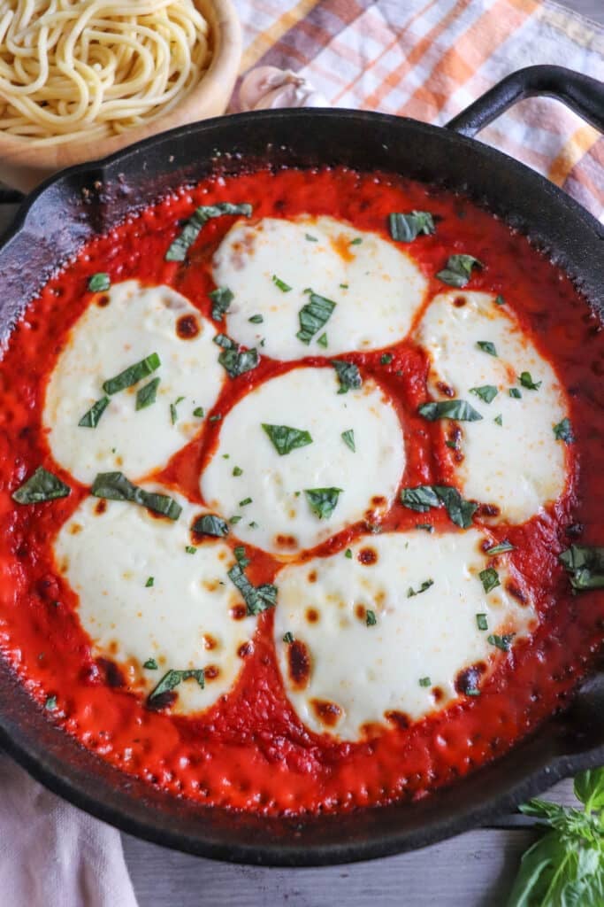 Mozzarella cheese slices on a bed of tomato sauce and chicken in a cast iron skillet.