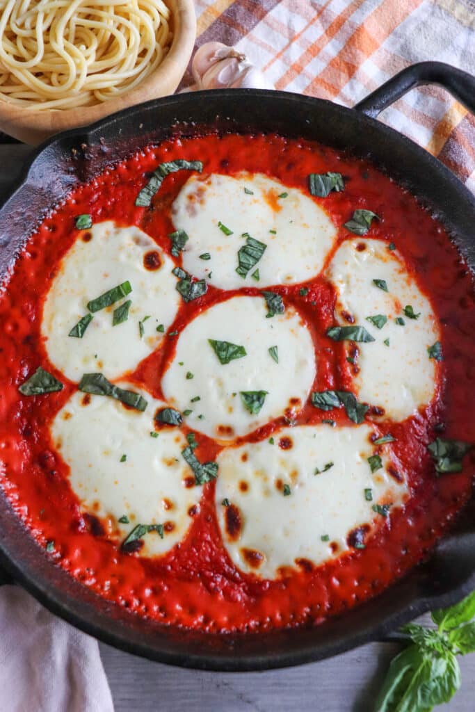 Mozzarella cheese slices on a bed of tomato sauce and chicken, garnished with fresh basil in a cast iron skillet.
