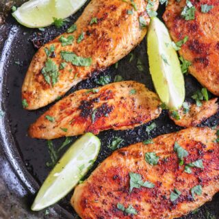 Blackened chicken breasts with lime wedges in a cast iron skillet with cilantro garnish.