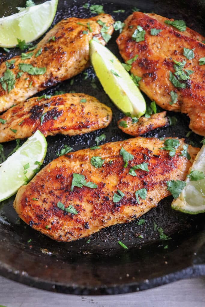 Close up of blackened chicken breasts with lime wedges in a cast iron skillet with cilantro garnish.