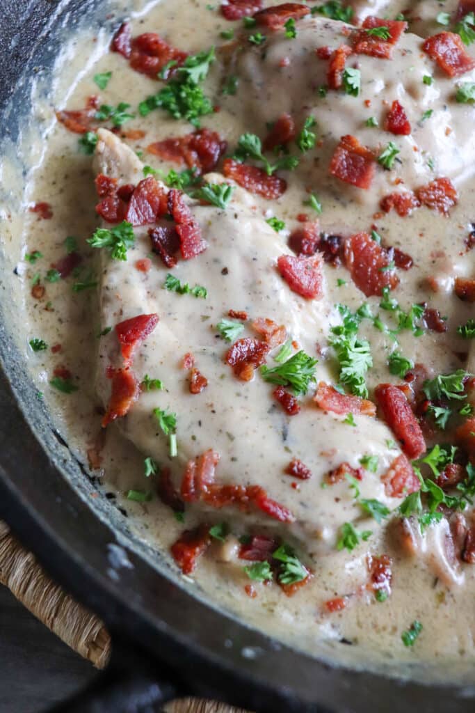 Close up of chicken in a cream sauce with bacon and parsley garnish in a cast iron skillet.