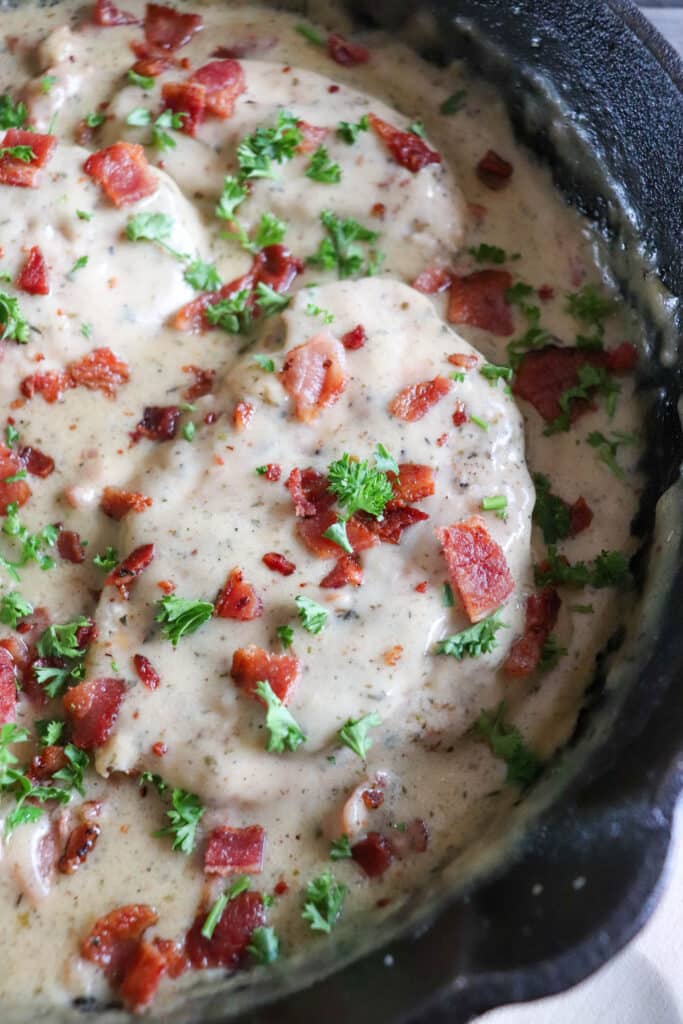 Close up of chicken in a cream sauce with bacon and parsley garnish in a cast iron skillet.