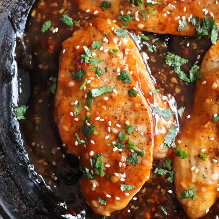 Close up of cooked chicken breast covered in sweet chili sauce, garnished with cilantro and sesame seeds in a cast iron skillet.