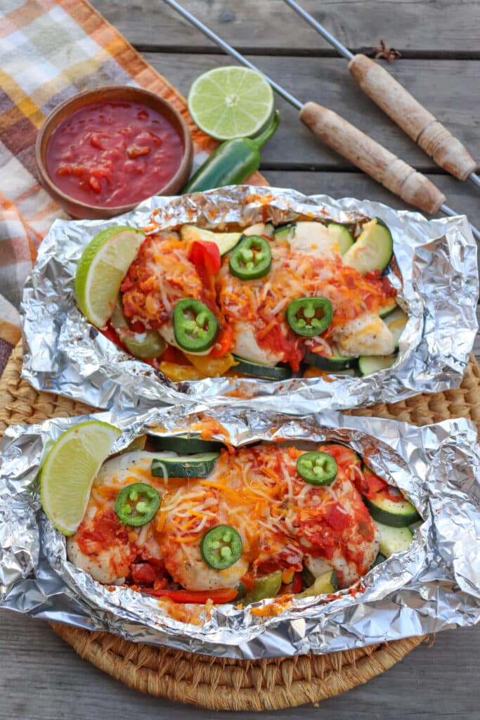 Salsa and cheese covering chicken in foil packets topped with slices of jalapeno and lime wedges on a wooden picnic table.