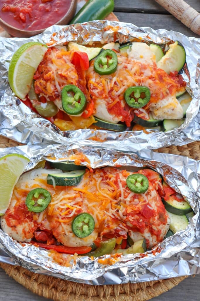 Salsa and cheese over chicken in foil packets with jalapeno slices and lime wedges on a picnic table.