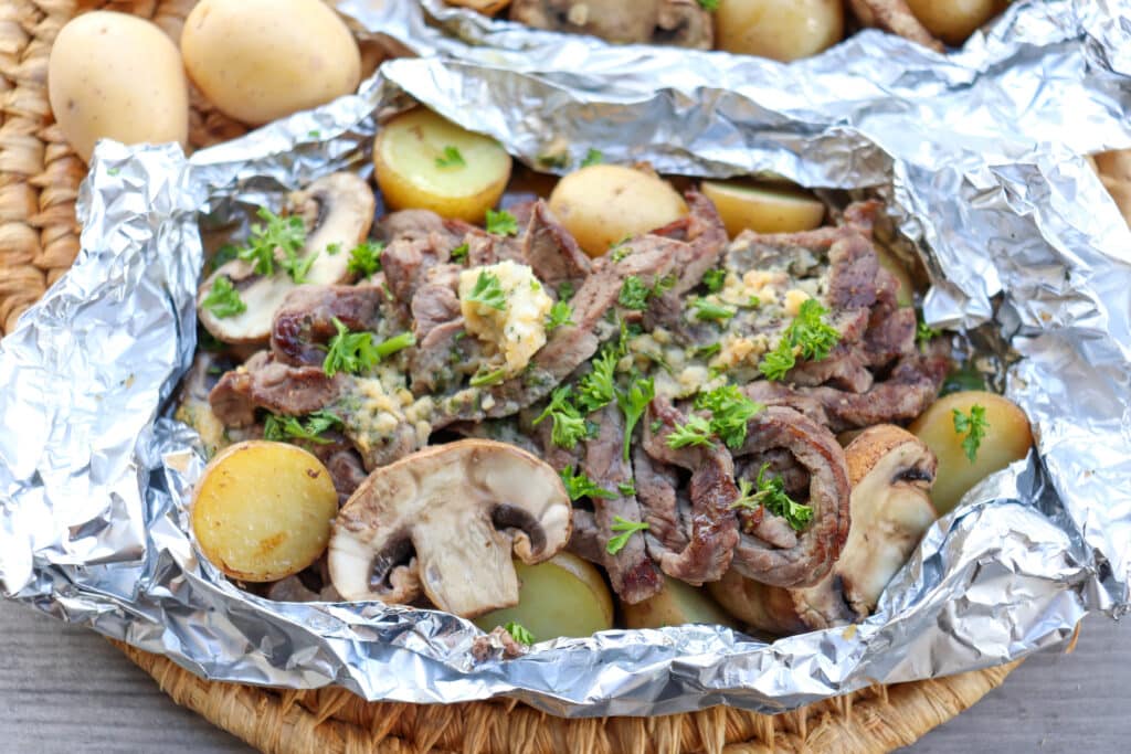 Steak, mushrooms and potatoes in a foil packet on a wooden picnic table.