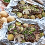 Steak, mushroom and potatoes in foil packets on a wooden picnic table.