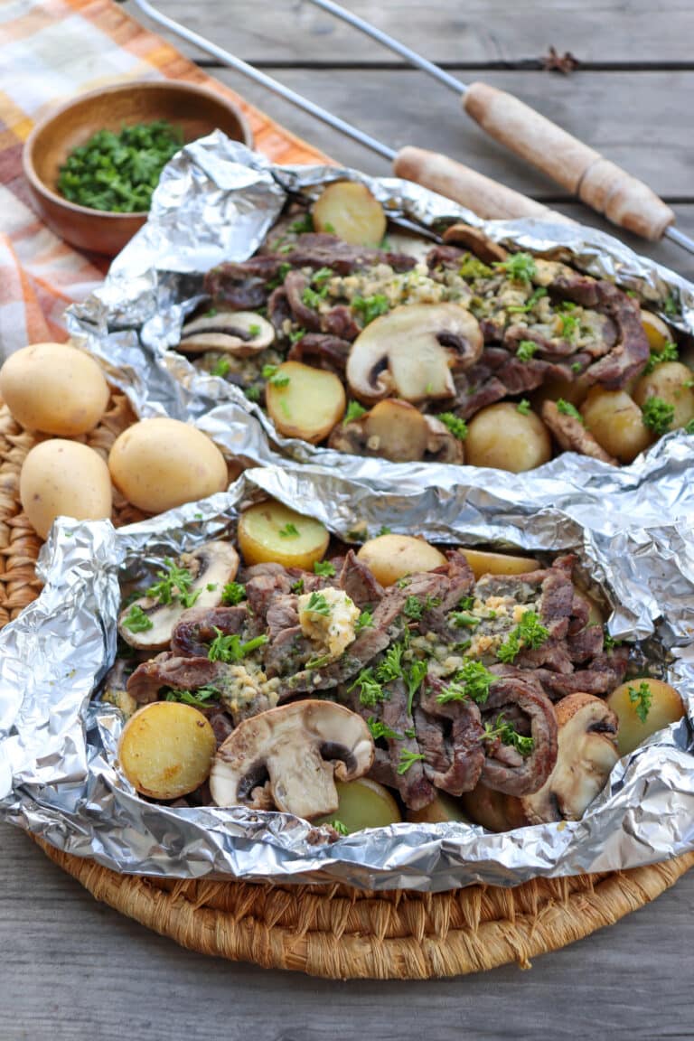 Steak, mushroom and potatoes in foil packets on a wooden picnic table.