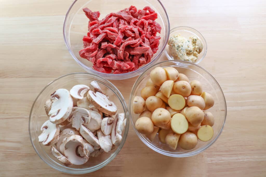 Ingredients for steak and mushroom foil packs in clear glass bowls on a wooden table.