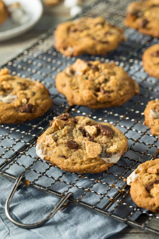 Freshly baked cookies with chocolate chips and marshmallow bits, reminiscent of a classic smores cookie recipe, are cooling on a wire rack covered with a blue cloth. A plate with more cookies is partially visible in the background.