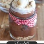 A jar of chocolate smores dessert with toasted marshmallow topping and a piece of chocolate. Its tied with a red gingham ribbon. The background includes another jar and crumbs. Text reads chocolate Smores Dessert CAMPFIREDOODIE.COM.