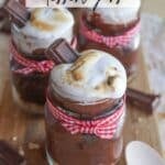 Smores dessert jars filled with layers of chocolate, graham cracker crumbs, and toasted marshmallow. Each jar is tied with a red gingham ribbon and topped with a chocolate square. A wooden spoon is visible nearby.