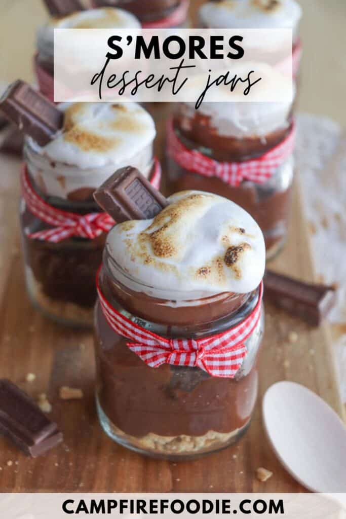 Smores dessert jars filled with layers of chocolate, graham cracker crumbs, and toasted marshmallow. Each jar is tied with a red gingham ribbon and topped with a chocolate square. A wooden spoon is visible nearby.