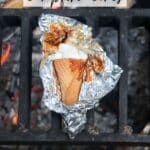 A waffle cone filled with marshmallows and chocolate, partially wrapped in foil, sits on a campfire grill over burning coals. Text reads Smores campfire cones and campfirefoodie.com.