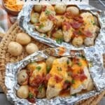 Two foil packets on a woven mat containing chicken, baby potatoes, melted cheese, bacon bits, and herbs. A small bowl of shredded cheese and a plaid cloth are beside them. Text on the image reads Foil Packet Chicken Ranch and campfirefoodie.com.