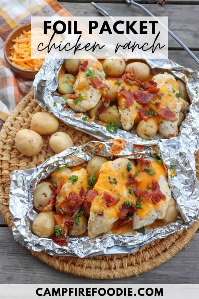 Two foil packets on a woven mat containing chicken, baby potatoes, melted cheese, bacon bits, and herbs. A small bowl of shredded cheese and a plaid cloth are beside them. Text on the image reads Foil Packet Chicken Ranch and campfirefoodie.com.