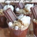 Close-up of several mini chocolate pudding cups topped with whipped cream, crumbled graham crackers, and small chocolate bars. Each cup has a few mini marshmallows. Text on image reads Smores pudding shots and campfirefoodie.com.