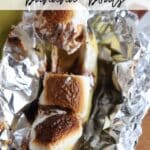 A banana split open and filled with toasted marshmallows and chocolate is nestled in a piece of foil. The text CAMPING banana boats is written above, and the website CAMPFIREEFOODIE.COM is displayed at the bottom.