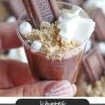 A hand holds a small dessert cup filled with chocolate pudding, topped with a piece of chocolate, whipped cream, graham cracker crumbs, and mini marshmallows. The overlay text reads Smores Pudding Shots and campfirefoodie.com.