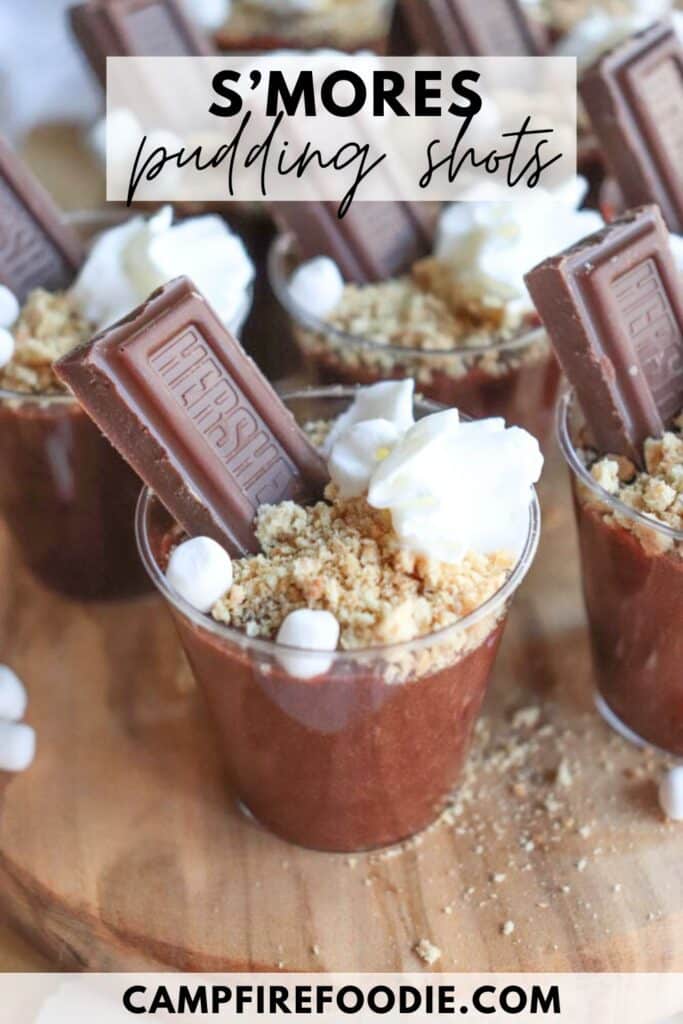 Clear cups filled with chocolate pudding, topped with crumbled graham crackers, mini marshmallows, whipped cream, and chocolate bars. Text on the image reads Smores pudding shots and campfirefoodie.com. Cups are arranged on a wooden surface.