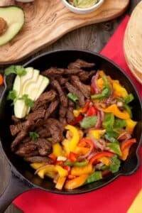 A skillet of Skillet Steak Fajitas brimming with sliced cooked beef, vibrant bell peppers, red onion, and cilantro. Avocado slices garnish the dish. In the background, a wooden board holds tortilla wraps and a partial view of a small dish with sauce.