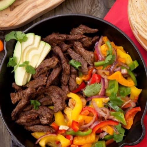 A skillet of Skillet Steak Fajitas brimming with sliced cooked beef, vibrant bell peppers, red onion, and cilantro. Avocado slices garnish the dish. In the background, a wooden board holds tortilla wraps and a partial view of a small dish with sauce.