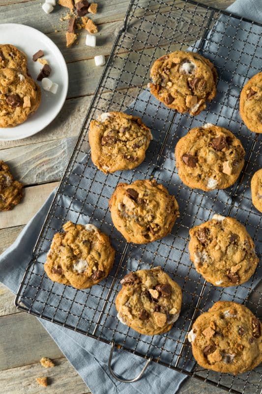 Homemade smores cookies with chocolate and marshmallow pieces rest on a cooling rack, placed over a blue cloth. A white plate with more cookies and scattered marshmallows adds charm to the wooden surface, making this smores cookie recipe a delightful treat.