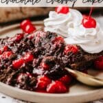 A close-up of a chocolate cherry cake topped with cherries and two dollops of whipped cream. The cake is on a plate with a spoon. Text on the image reads Dutch Oven Chocolate Cherry Cake and campfirefoodie.com.