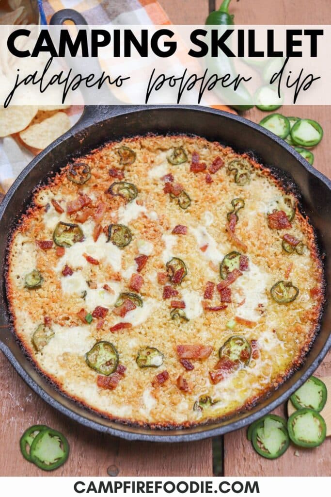 A skillet filled with jalapeño popper dip, topped with sliced jalapeños and bits of bacon. Its set on a wooden surface with jalapeño slices and a checkered cloth nearby. Text reads: Camping Skillet jalapeno popper dip.