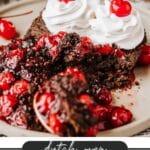 A chocolate cherry cake topped with whipped cream and cherries. The cake is sliced and served on a plate, with cherry filling oozing out. Text at the bottom reads Dutch Oven Cherry Cake - CampfireFoodie.com.