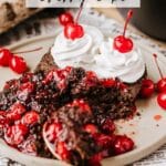 A slice of Dutch oven cherry cake topped with cherries and whipped cream on a plate. A spoon rests beside the cake, surrounded by more cherries, with a piece of log and a black pot in the background. The text reads Dutch Oven Cherry Cake.