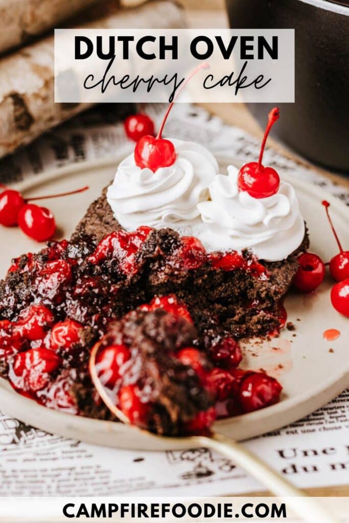 A slice of Dutch oven cherry cake topped with cherries and whipped cream on a plate. A spoon rests beside the cake, surrounded by more cherries, with a piece of log and a black pot in the background. The text reads Dutch Oven Cherry Cake.