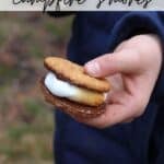 A person in a blue jacket holds a freshly made smore with toasted marshmallow and melted chocolate between two graham crackers. Text at the top says, MAKE THE BEST campfire s’mores, and at the bottom, CAMPFIREEFOODIE.COM.