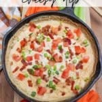 A skillet filled with creamy, cheesy dip topped with diced tomatoes, bacon bits, and green onions. Surrounded by carrot and red bell pepper sticks on a wooden surface. Text overlay reads Skillet Cheesy Dip and campfirefoodie.com.