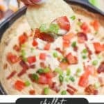 A hand holds a chip dipped in a creamy skillet cheesy dip, topped with chopped tomatoes, green onions, and bacon. Text at the bottom reads, Skillet Cheesy Dip, campfirefoodie.com.