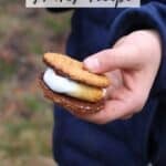 A person in a dark blue jacket holds a smore with melted chocolate and marshmallow between graham crackers. Text reads The Best Smores Recipe and campfirefoodie.com at the bottom.