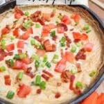 A skillet filled with cheesy dip topped with chopped tomatoes, green onions, and crispy bacon pieces. The skillet is on a wooden surface with an orange-striped cloth partially visible. Text reads Skillet cheesy dip and campfirefoodie.com.