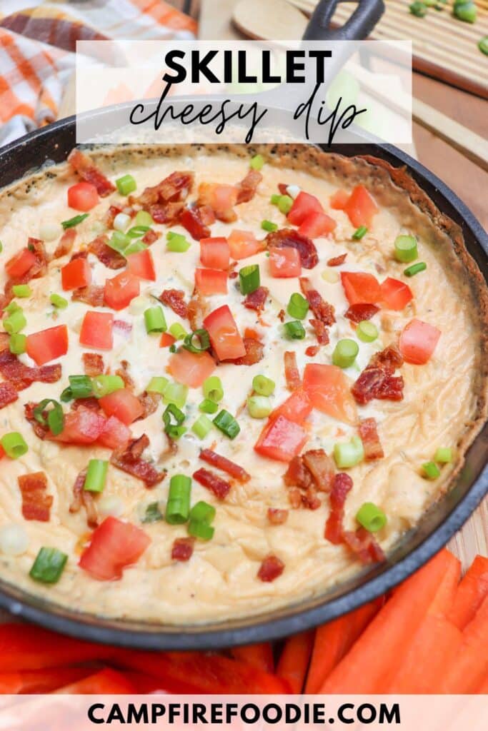 A skillet filled with cheesy dip topped with chopped tomatoes, green onions, and crispy bacon pieces. The skillet is on a wooden surface with an orange-striped cloth partially visible. Text reads Skillet cheesy dip and campfirefoodie.com.