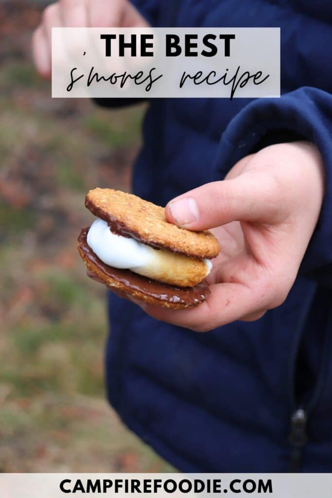 A person in a dark blue jacket holds a smore with melted chocolate and marshmallow between graham crackers. Text reads The Best Smores Recipe and campfirefoodie.com at the bottom.