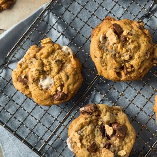 A freshly baked batch of smores cookies with chocolate chunks and marshmallows sits cooling on a wire rack, perfecting this recipe. The rack rests on a blue cloth atop a wooden surface, while one cookie, a masterpiece in itself, is displayed separately in the background.