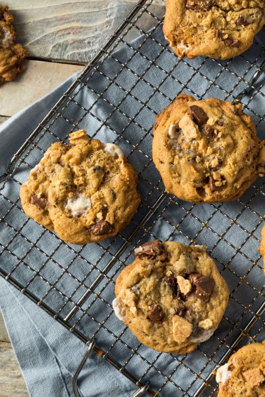 A freshly baked batch of smores cookies with chocolate chunks and marshmallows sits cooling on a wire rack, perfecting this recipe. The rack rests on a blue cloth atop a wooden surface, while one cookie, a masterpiece in itself, is displayed separately in the background.