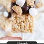 A rice krispies treat on a stick, topped with chocolate, crushed graham crackers, and mini marshmallows. The text on the image reads Smores Rice Krispies and campfirefoodie.com at the bottom.