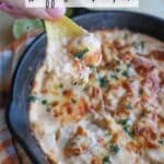 A person holds a chip topped with melted buffalo chicken dip, featuring cheese and herbs, over a sizzling skillet of the same dip. The skillet is on a wooden surface, and theres a text overlay that reads Skillet Dip Buffalo Chicken.