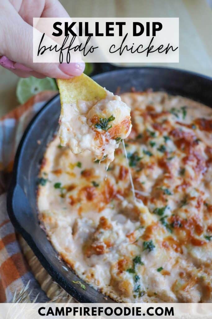 A person holds a chip topped with melted buffalo chicken dip, featuring cheese and herbs, over a sizzling skillet of the same dip. The skillet is on a wooden surface, and theres a text overlay that reads Skillet Dip Buffalo Chicken.