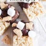 Close-up of smores rice krispies treats on sticks. Topped with chocolate, mini marshmallows, chocolate chips, and graham cracker pieces. Placed on white parchment paper with text overlay: Smores rice krispies and campfirefoodie.com.