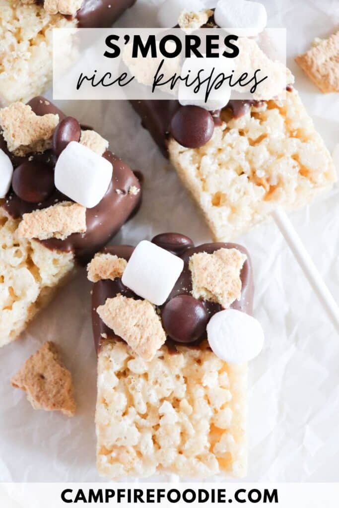 Close-up of smores rice krispies treats on sticks. Topped with chocolate, mini marshmallows, chocolate chips, and graham cracker pieces. Placed on white parchment paper with text overlay: Smores rice krispies and campfirefoodie.com.