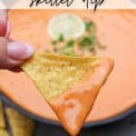 A close-up of a hand holding a triangular tortilla chip dipped in creamy queso skillet dip. The dip is smooth and orange with a garnish in the background. Text at the top reads QUESO skillet dip, and the bottom text says CAMPFIREDOODIE.COM.