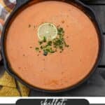 A skillet filled with creamy queso dip garnished with a lime slice and chopped green peppers. Next to the skillet are tortilla chips and a sliced lime on a checkered cloth. Text below reads Skillet Queso Dip from campfirefoodie.com.