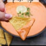 A hand holds a tortilla chip dipped in creamy orange skillet queso, garnished with a lime slice and herbs. In the background, a bowl of chips is visible. Text on the image reads Skillet queso dip and campfirefoodie.com.